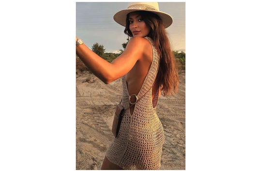 Mulher usando vestido boho tricotado natural com costas à mostra, chapéu de palha, em cenário de praia ao pôr do sol - moda verão 2025/2026