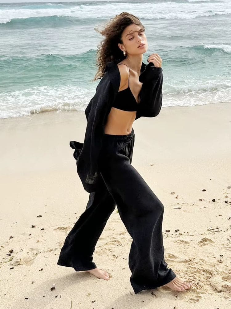 Modelo caminhando descalça na praia vestindo o conjunto de linho preto com camisa aberta em um estilo balneário despojado
